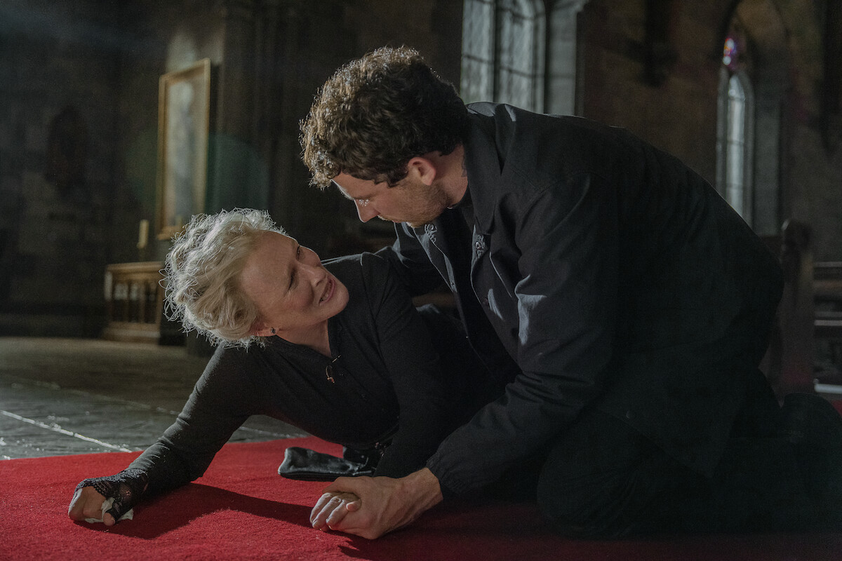 An older woman on the floor of a dimly lit church looks up in distress at a man leaning over her. The mood is tense and dramatic, with emotional intensity and gothic atmosphere in the historic setting.