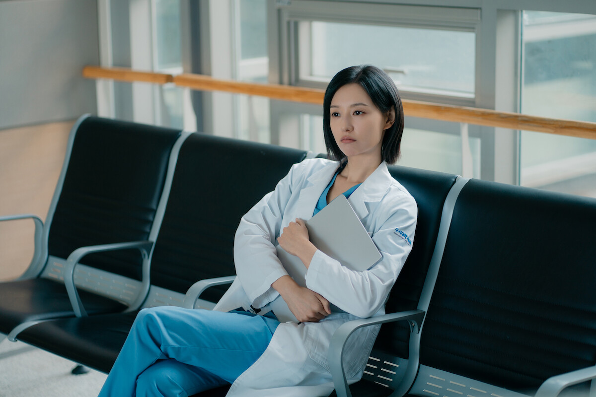 Uma mulher asiática de cabelos pretos está sentada no banco de espera de um hospital segurando uma prancheta e vestindo jaleco branco