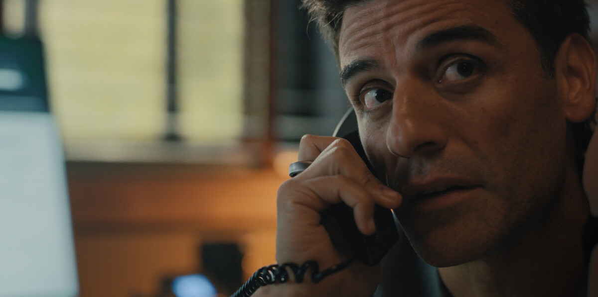 A man with a serious expression holds a phone to his ear indoors, with a blurred computer screen and window in the background, suggesting an office or work environment and a moment of intense conversation or concern.