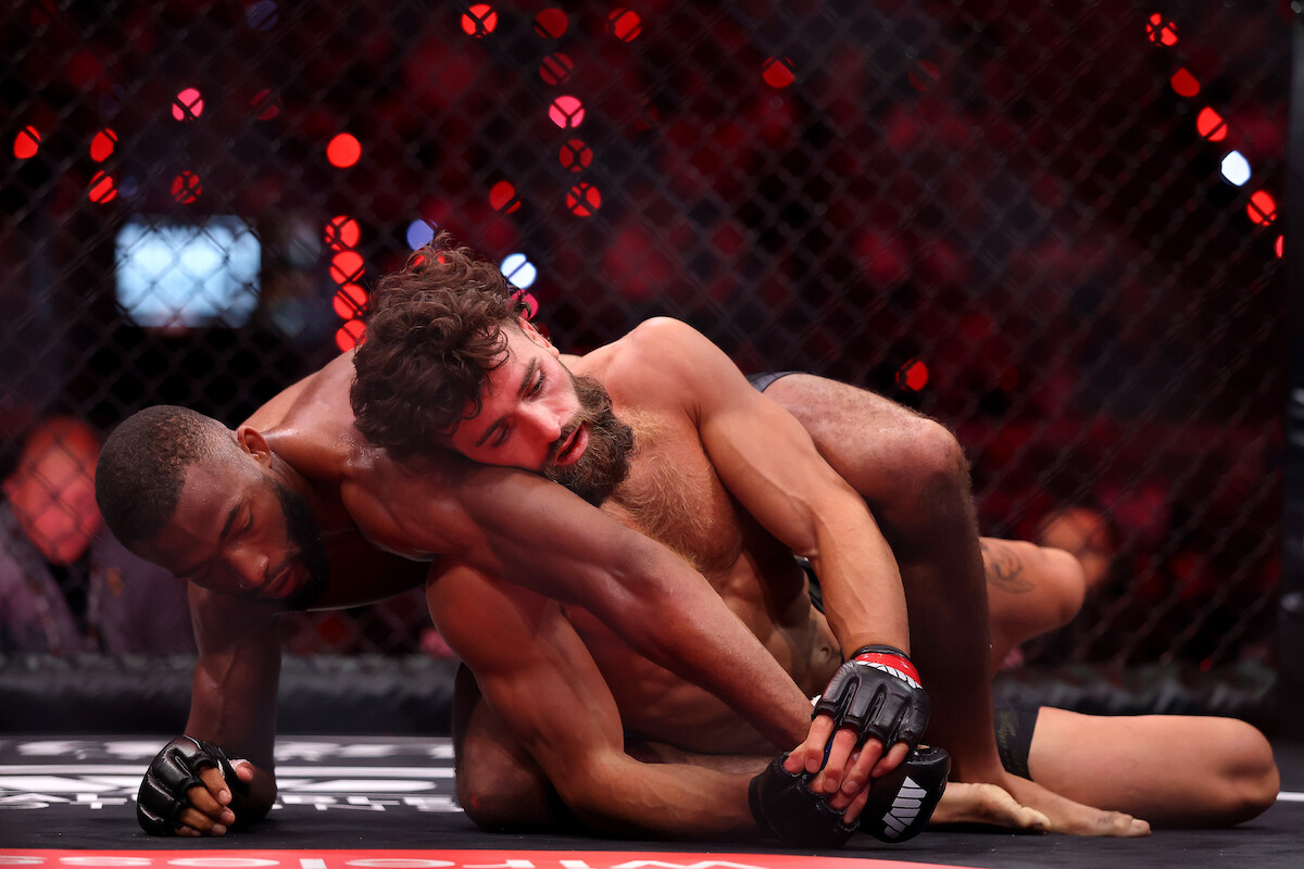 Two mixed martial artists grapple on the mat inside a caged octagon during a professional MMA match, with the crowd and cage visible in the background under arena lighting.