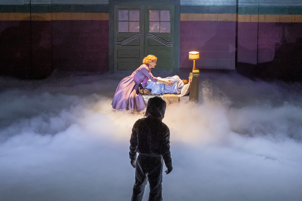 A person in a hooded outfit stands in fog watching a woman tend to someone lying in bed, set under dramatic stage lighting near a lamp and a closed double door, suggesting a theatrical or dreamlike scene.