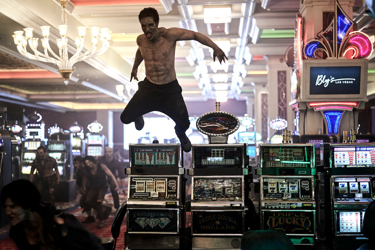 Shirtless man jumping over slot machines in a brightly lit casino with chandeliers, surrounded by slot machines, other people, and a Las Vegas sign in the background.