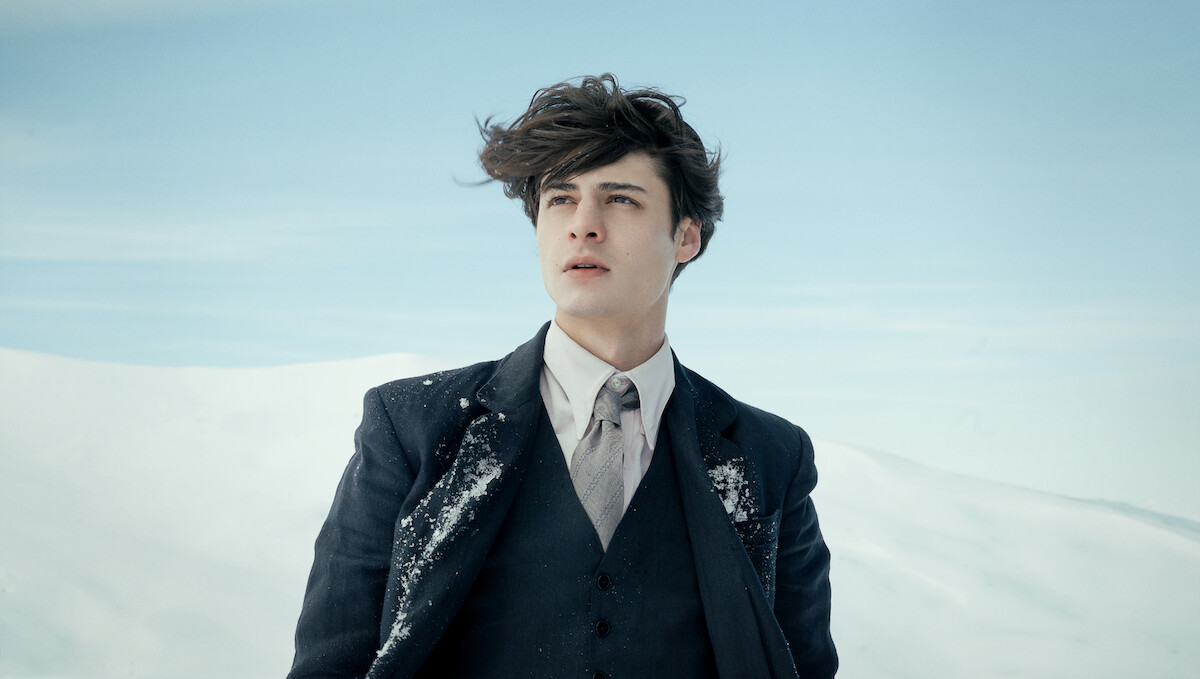 Young man in a suit stands in a snowy landscape with snow on his jacket, sky and snow-covered hills in the background, wind blowing his hair.
