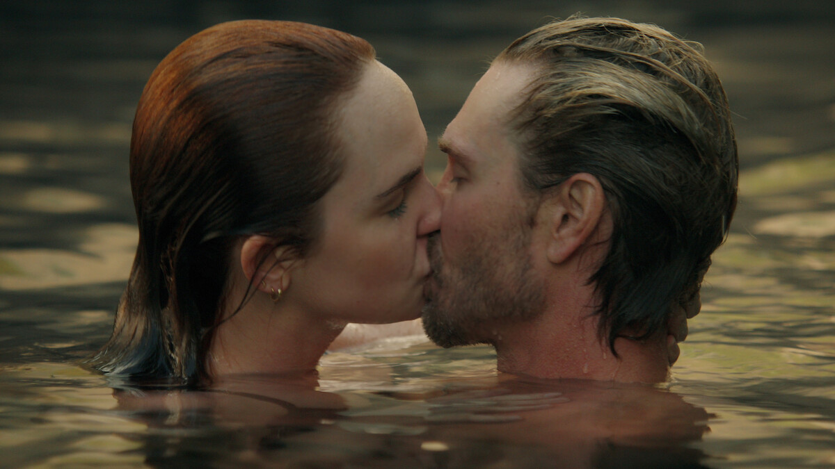 A man and a woman kissing in a body of water, both are partially submerged, with natural lighting and a serene outdoor environment.