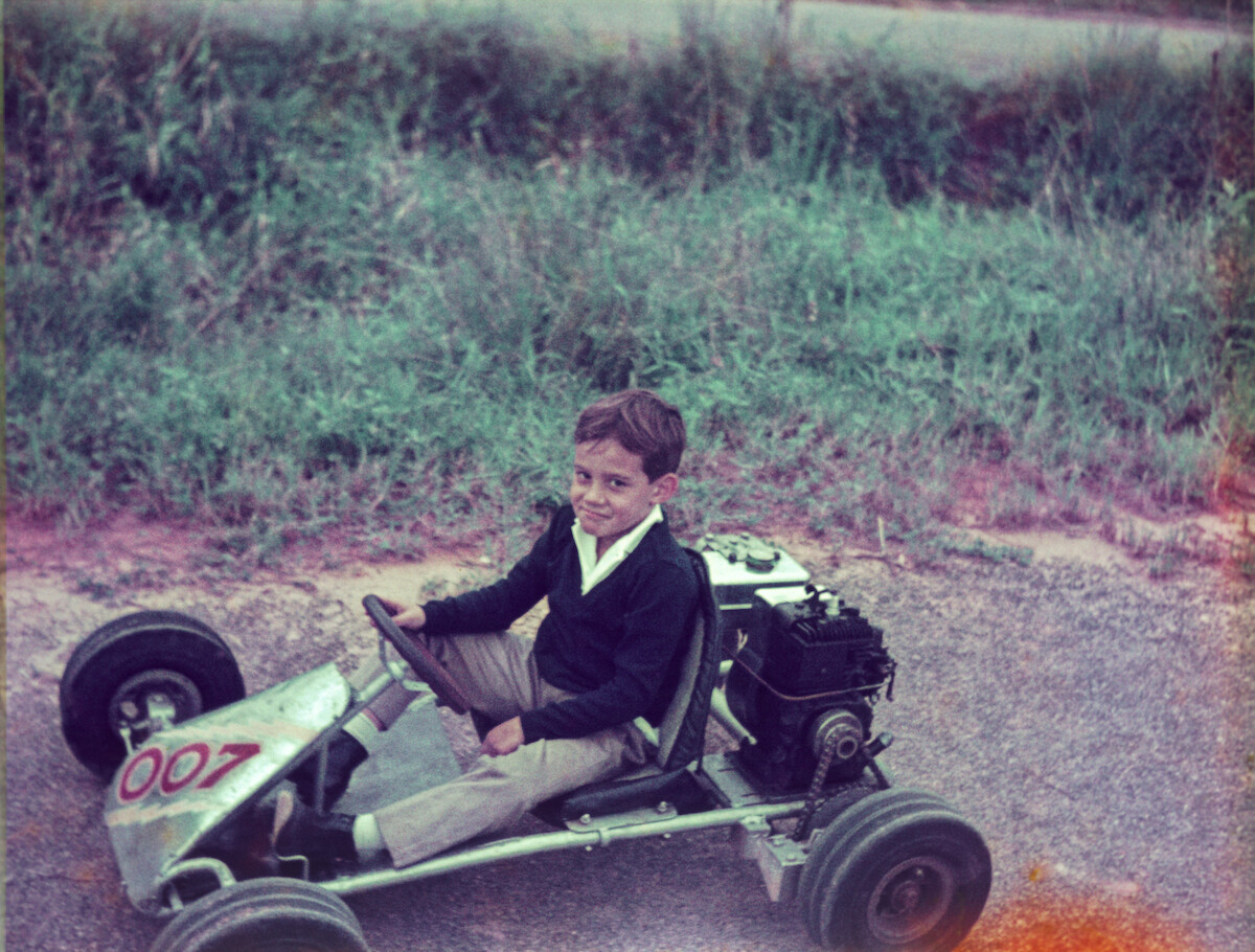 A very young Ayrton Senna wears a black top and races a silver car on a dirt road.