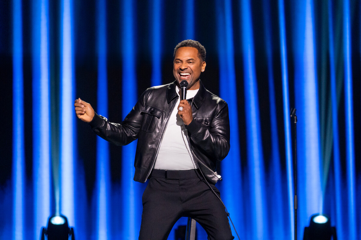 A comedian in a black leather jacket performs on stage with a microphone, smiling and animated, against a blue curtain backdrop with stage lights.