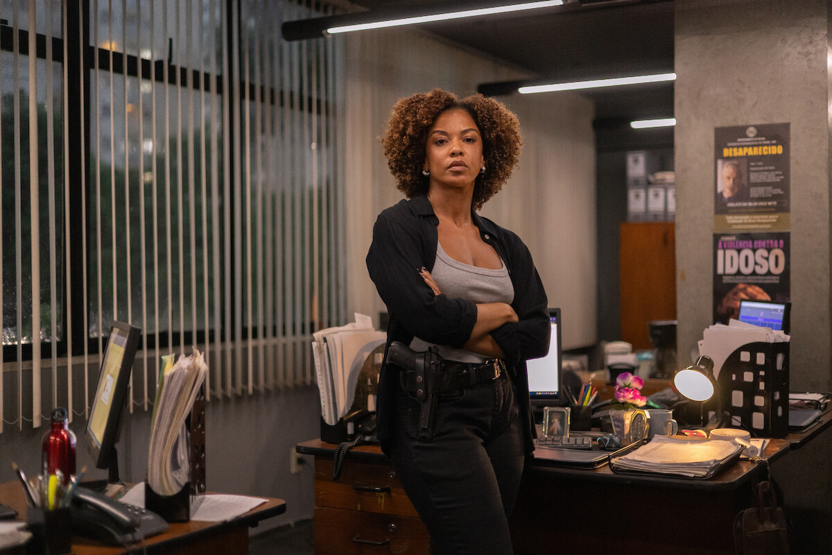 Uma policial negra, com cabelos volumosos e castanhos, está de pé em frente a uma mesa de uma delegacia