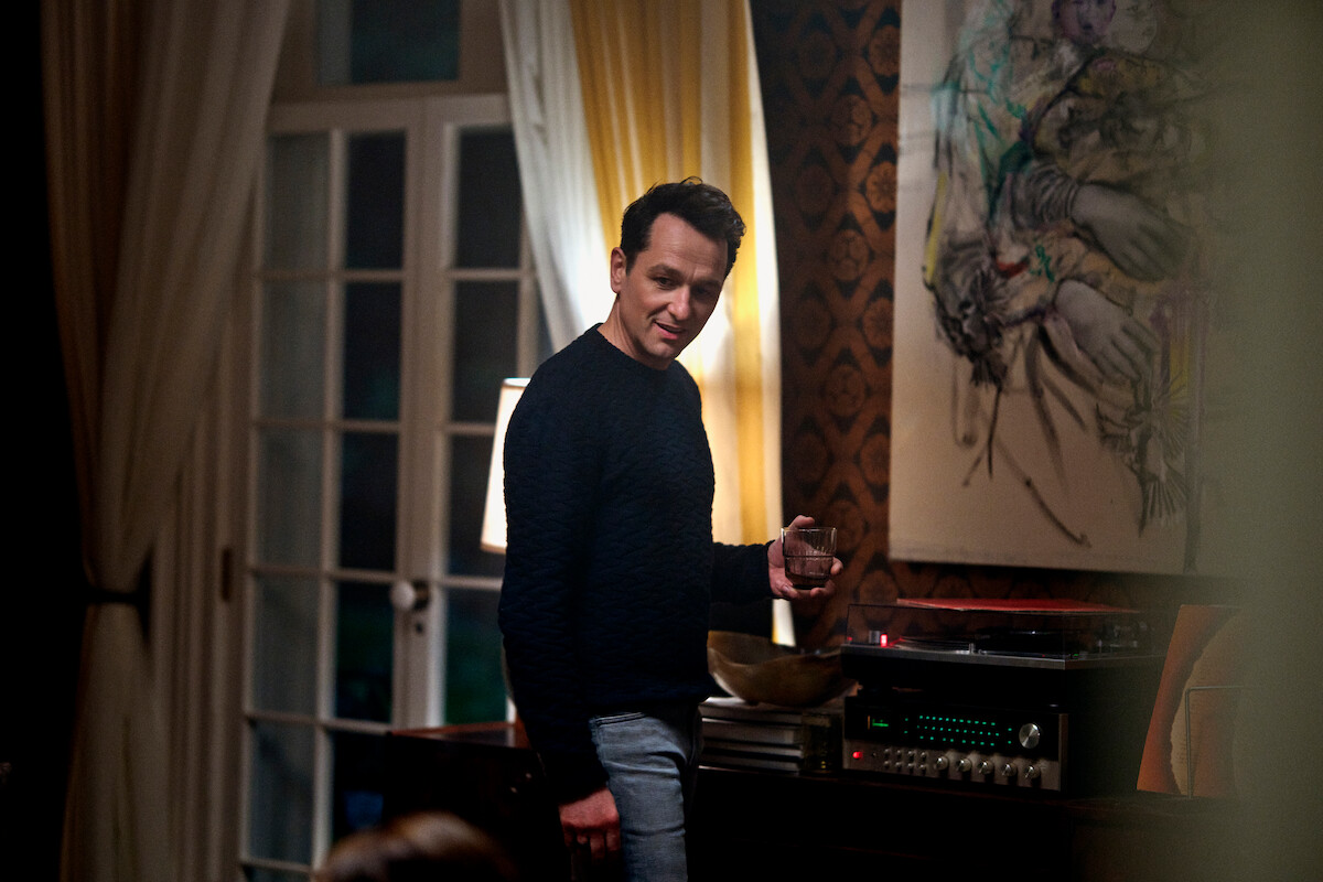 A man stands by a record player in a warmly lit, stylish living room with art on the wall and large windows, holding a drink, giving the scene a cozy and relaxed evening mood.