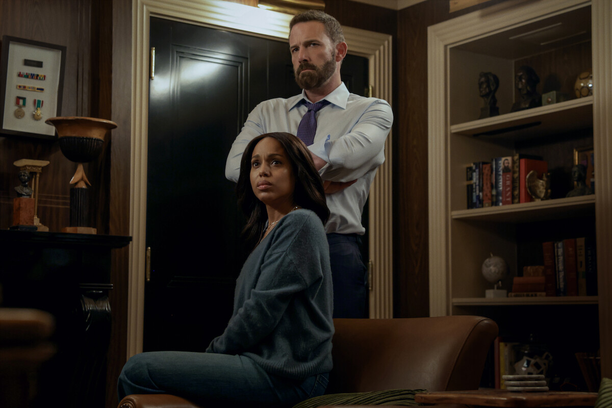 A woman sits on a leather chair looking concerned, while a man stands behind her with arms crossed in a dimly lit office or library filled with shelves, books, and decorative objects.