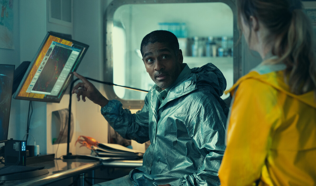 Man in a silver hazmat suit points at a computer monitor while talking to a woman in a yellow raincoat inside a laboratory or research station with scientific equipment and papers on the desk.