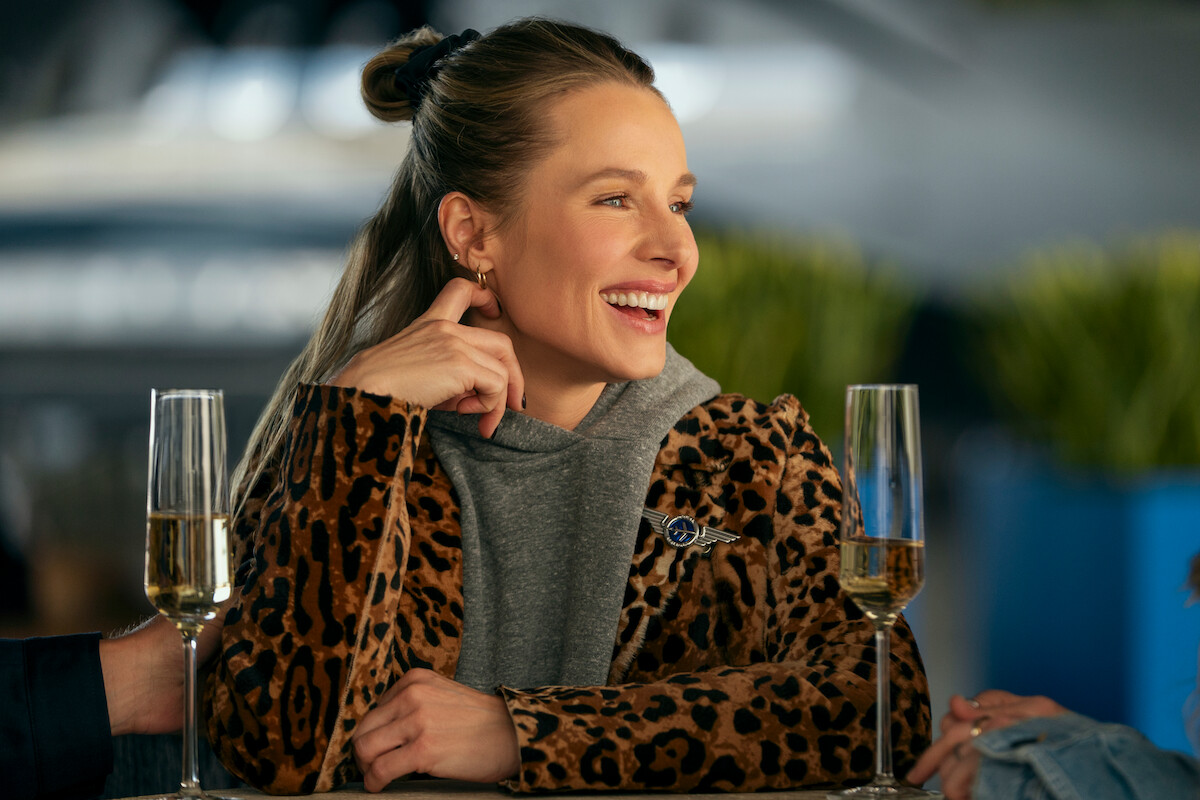 Woman in leopard print jacket sitting at a table, smiling and talking with others, two glasses of champagne in front of her, modern indoor setting with blurred background and plants.