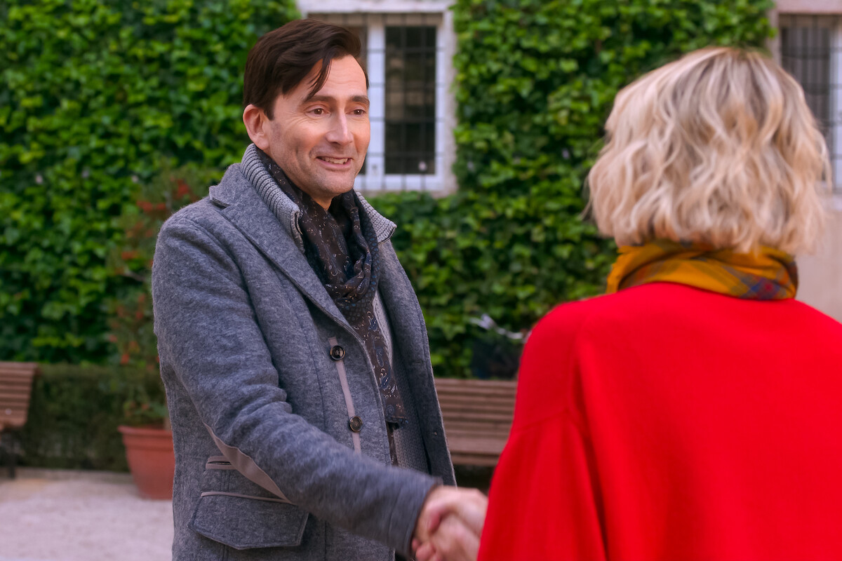 Man and woman shaking hands outdoors in a courtyard with greenery and benches, both wearing coats, smiling and engaged in conversation.
