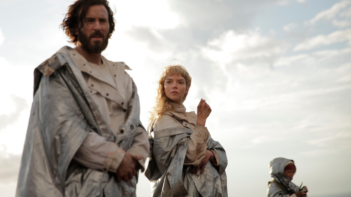 Three people in silvery cloaks stand outdoors under a cloudy sky, appearing serious and focused, with soft sunlight illuminating the scene.