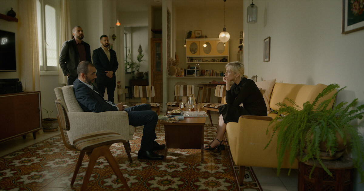 Four people in a living room with mid-century decor; two seated and talking, two standing behind, surrounded by vintage furniture, patterned floor, plants, and warm lighting.