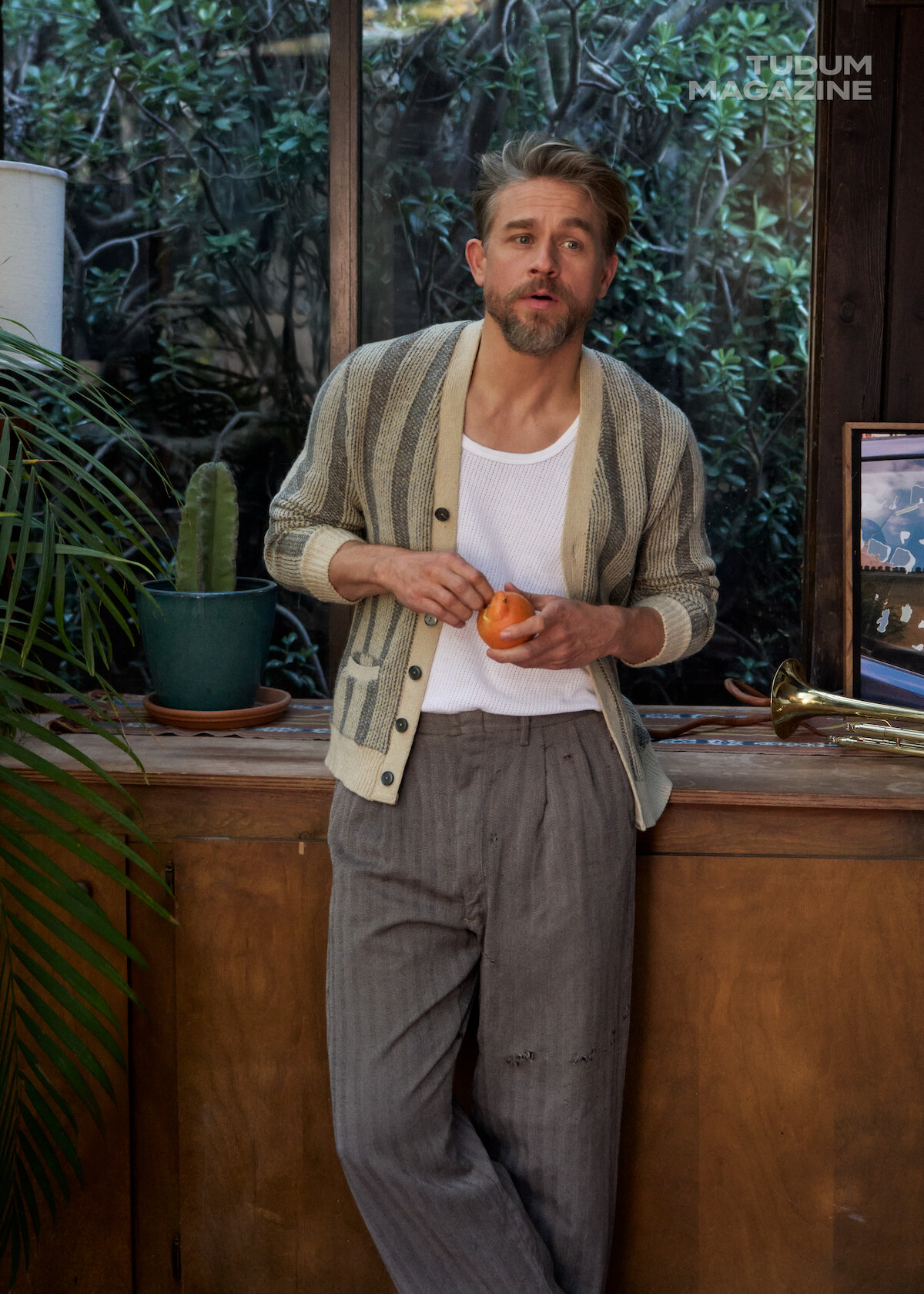 Charlie Hunnam leans against a cabinet in front of windows holding an orange that he is beginning to peel.