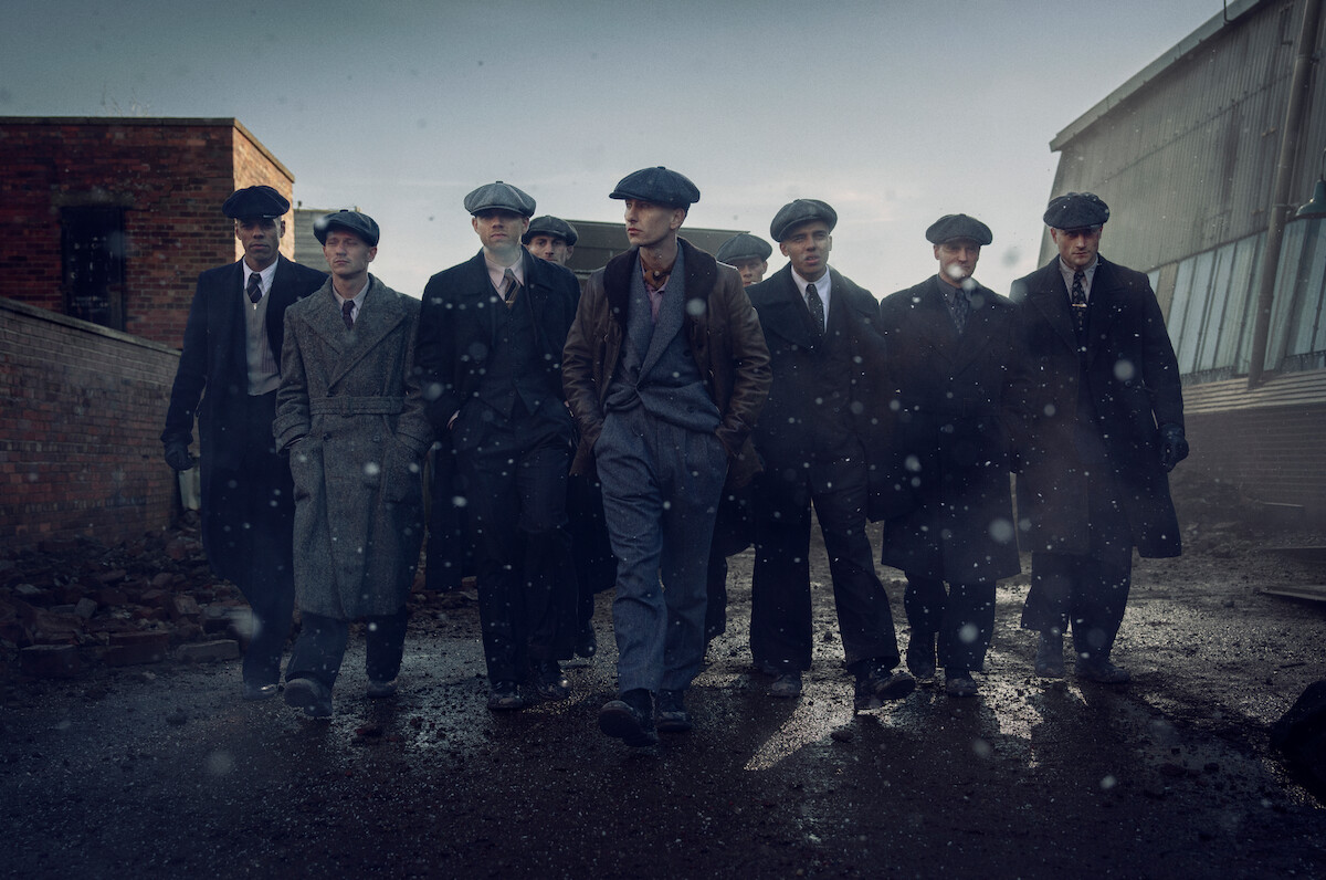 A group of men in vintage suits and newsboy caps walk together closely on a rainy, industrial street. The mood is serious and gritty, with cold light and falling rain enhancing the dramatic, early 20th-century atmosphere.