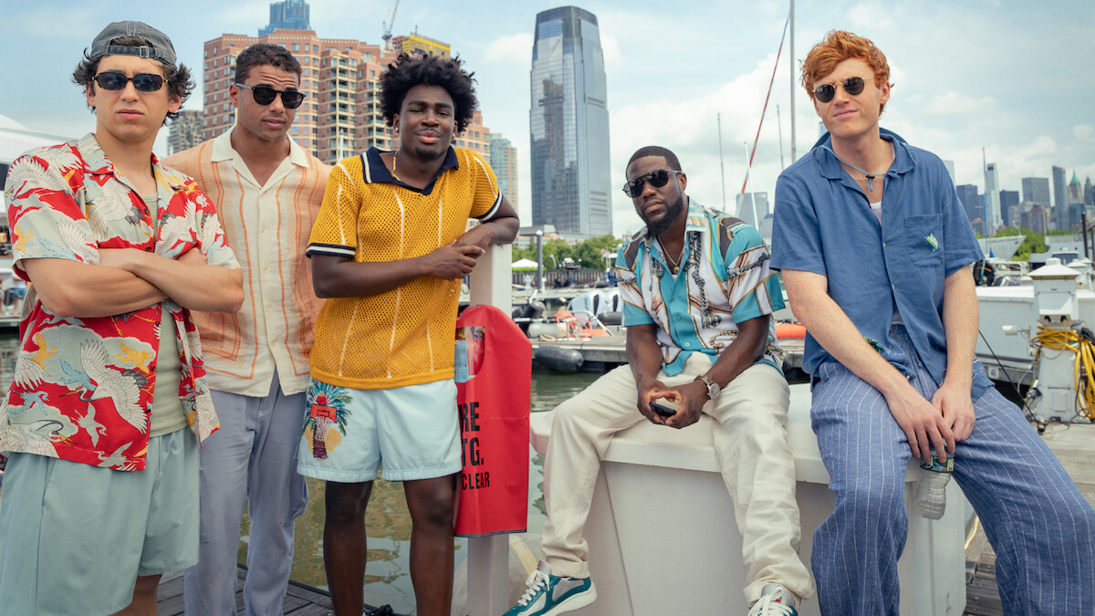 Five young men in casual summer outfits stand by a marina with boats, modern city buildings, and a waterfront skyline in the background, suggesting a relaxed and urban environment.