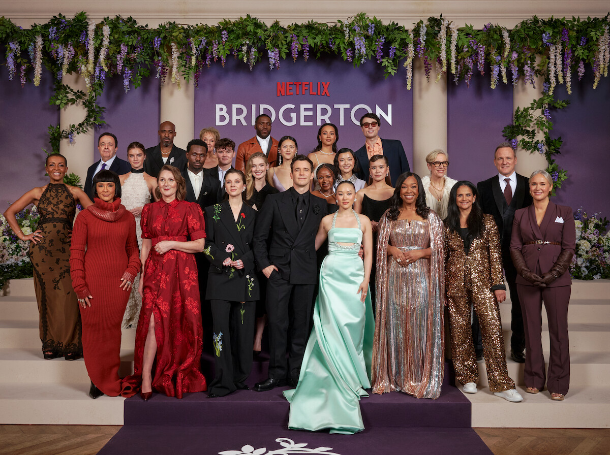 A group of elegantly dressed people posing on steps at a formal Netflix Bridgerton event, with floral decorations, purple backdrop, and lush greenery in the background.