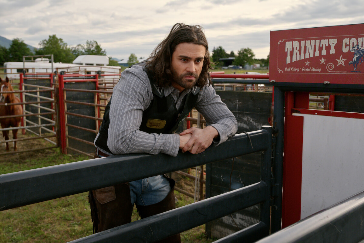 A man with long hair and a beard leans on a metal fence at a rodeo arena, wearing a striped shirt and vest. Horses, grass, and trailers appear in the background under a cloudy sky near signage reading "Trinity."