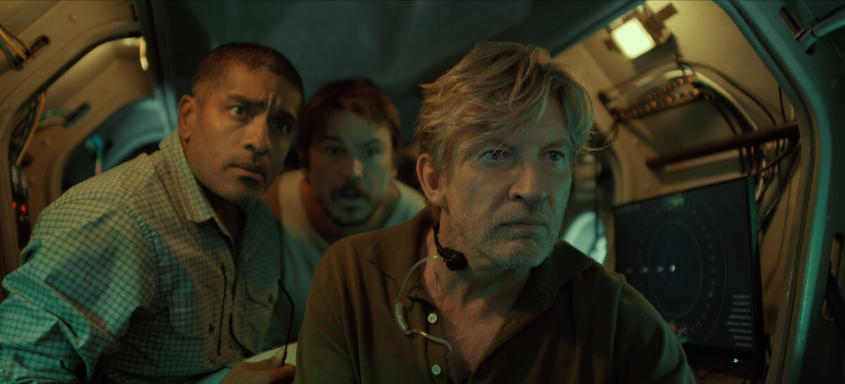 Three men in a dimly lit submarine interior look intently at a screen, surrounded by equipment and controls, suggesting a tense or serious moment in an underwater or scientific environment.