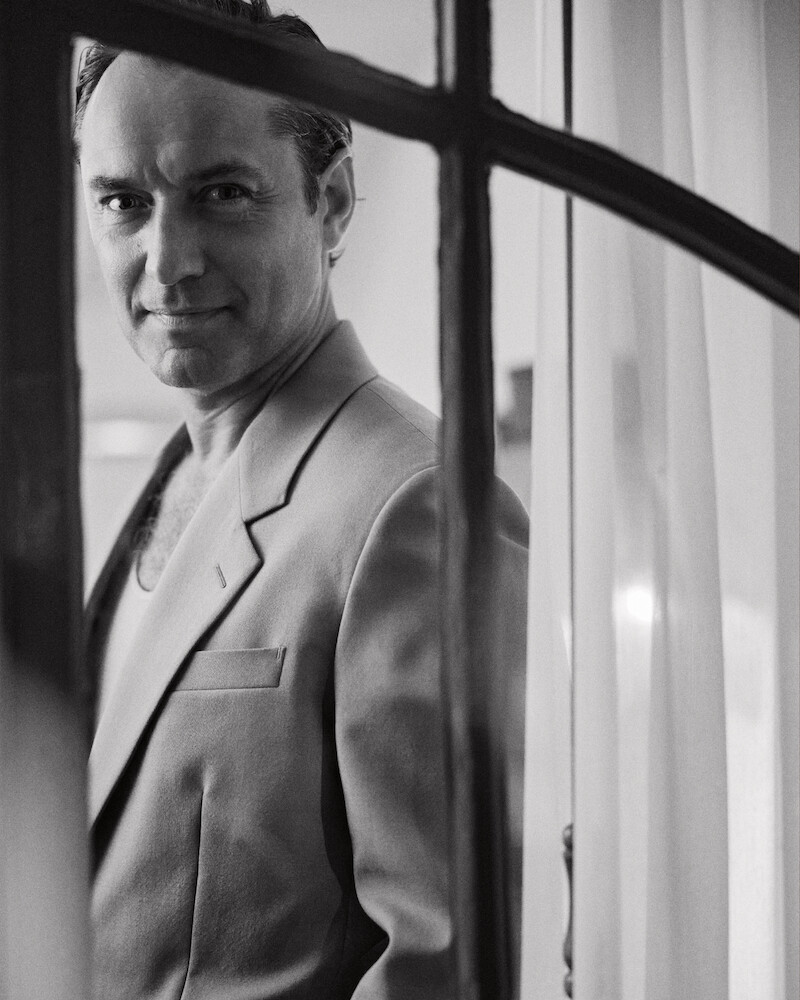 Man in a suit stands indoors by a window with sheer curtains, looking at the camera, captured in black and white with soft natural lighting.