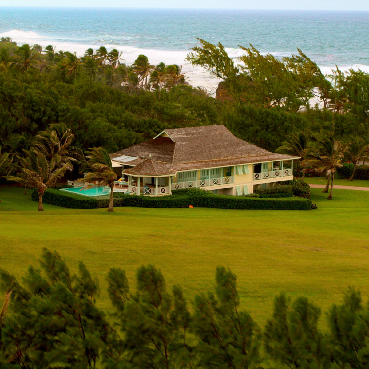A beachside house with a thatched roof surrounded by trees and lush green lawn, with a swimming pool nearby and the ocean visible in the background on a windy day.