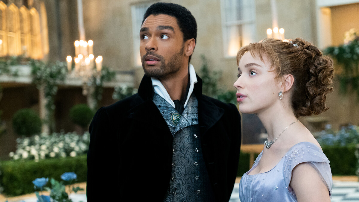 A well-dressed man and woman stand together in an elegant, candlelit ballroom or garden setting, surrounded by flowers and classical decor, looking upward with thoughtful expressions.