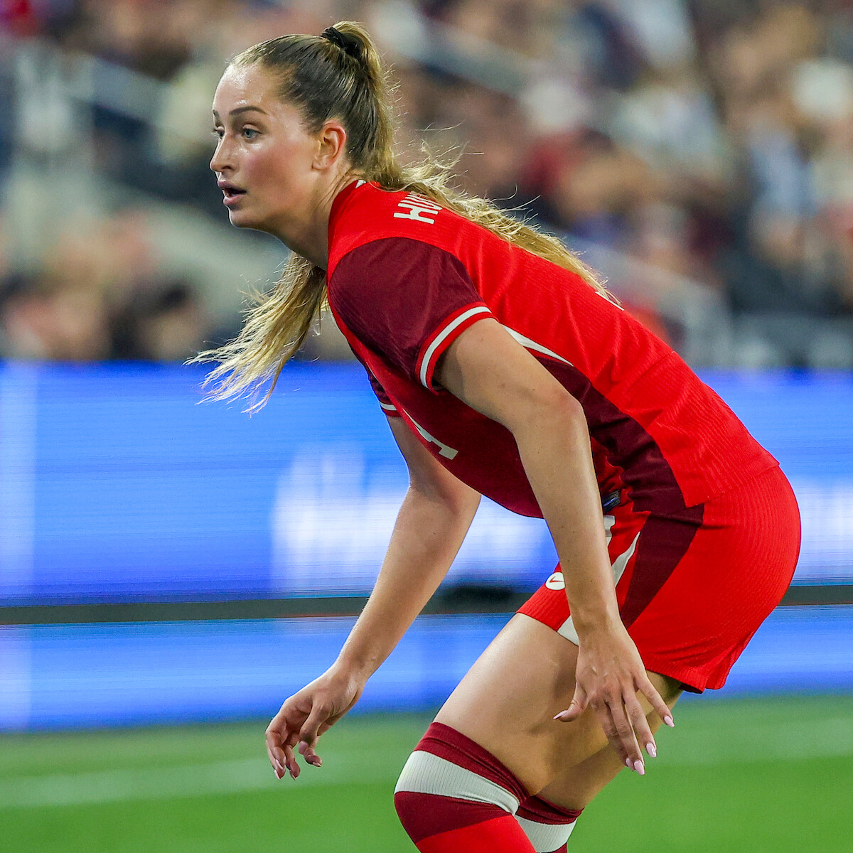 Female soccer player in a red uniform on a field during a match, with a blurred stadium crowd and LED displays in the background, focused and ready to play.