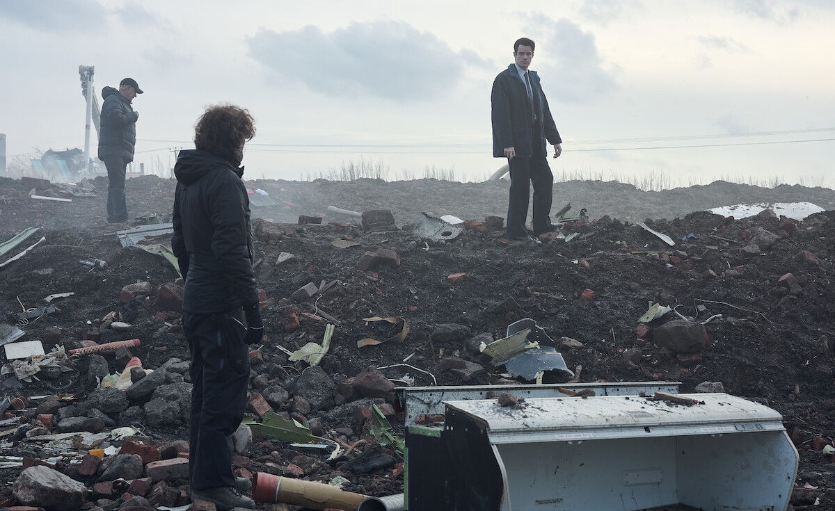 Three people inspecting debris and rubble on a desolate, cloudy landscape; two stand among scattered wreckage and broken objects, while one observes from a distance, suggesting a post-disaster or crime scene investigation setting.