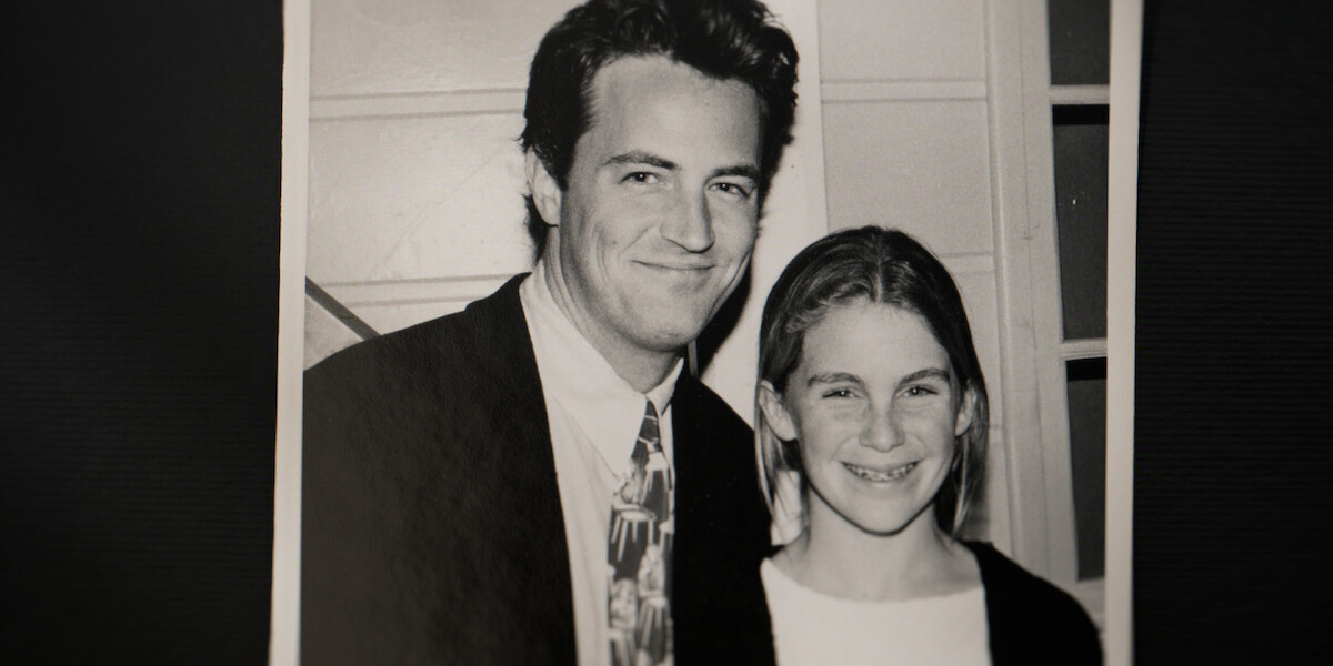 A black-and-white photo showing a man in a suit and tie smiling next to a young girl with braces. They are indoors, in front of a white wall with window panes. The atmosphere appears warm and friendly.