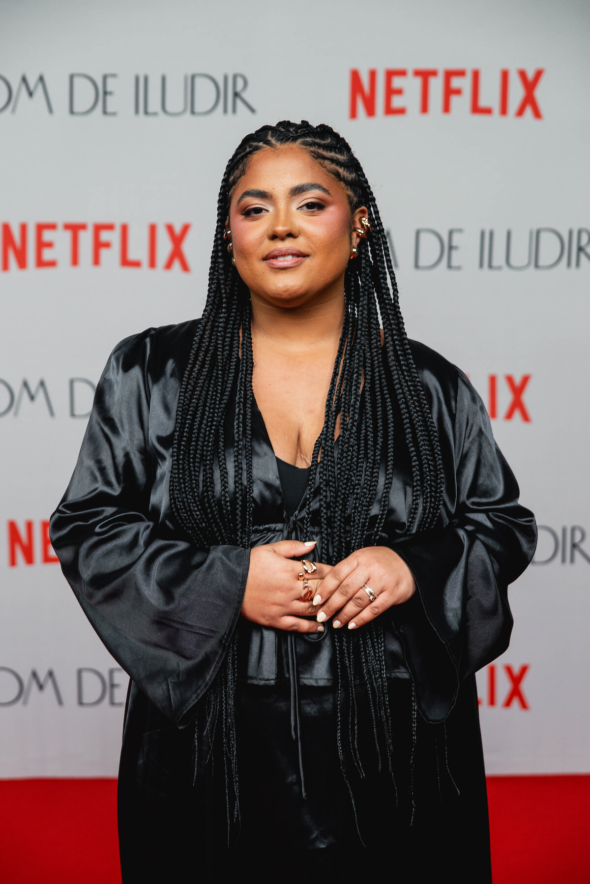 A atriz Giovanna de Jesus é uma mulher negra, com tranças nos cabelos e veste um vestido totalmente preto