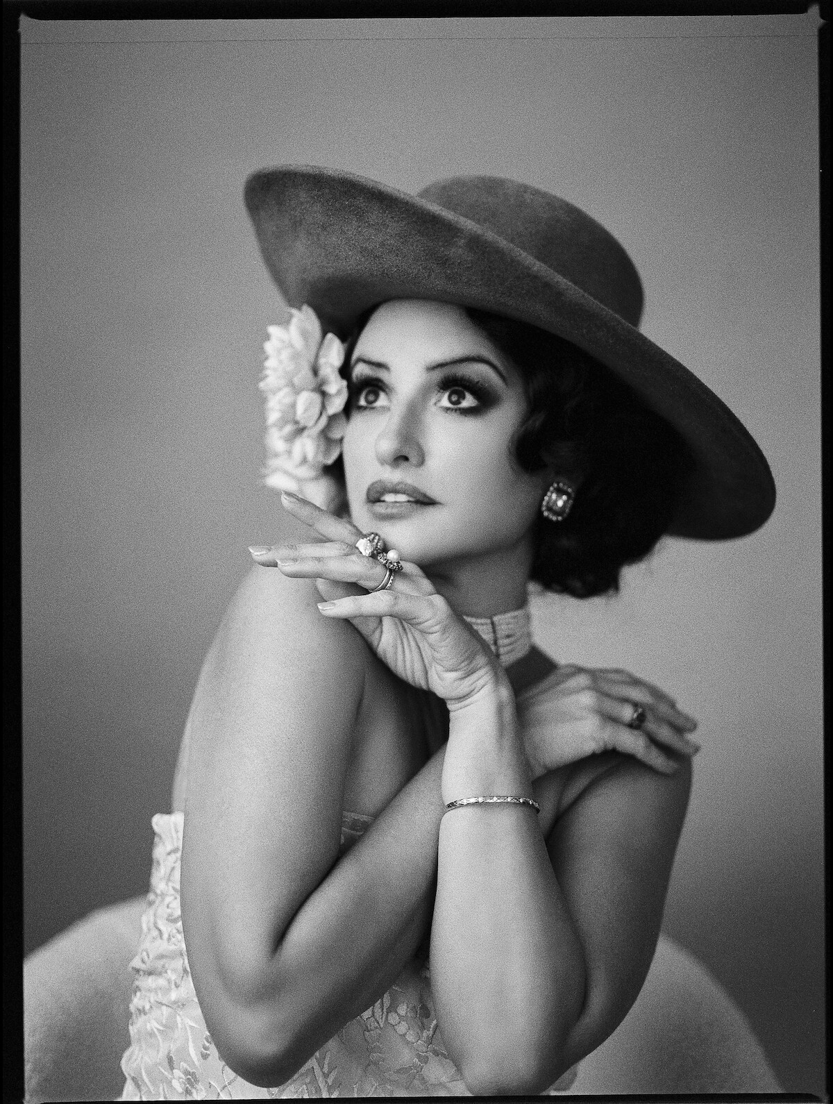 Penélope Cruz as Nené Romero in La Bola Negra in a black and white photograph wearing a strapless white lace dress with a choker necklace made of pearls. She also has a wide-brimmed velvet hat on with a flower tucked behind her ear while looking up with one hand positioned under her chin and another on her shoulder.