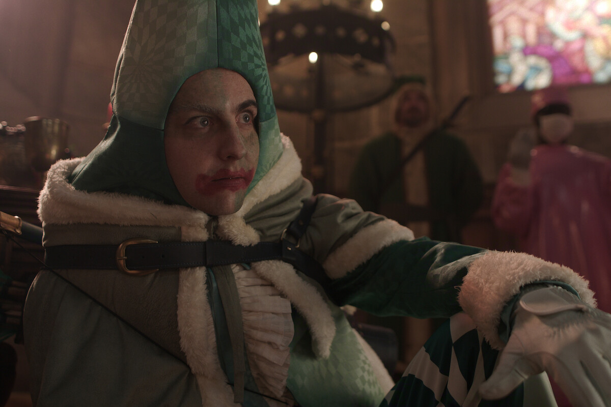 A jester in green and white costume sits indoors in a medieval-style room with stained glass and people in historical attire in the background.