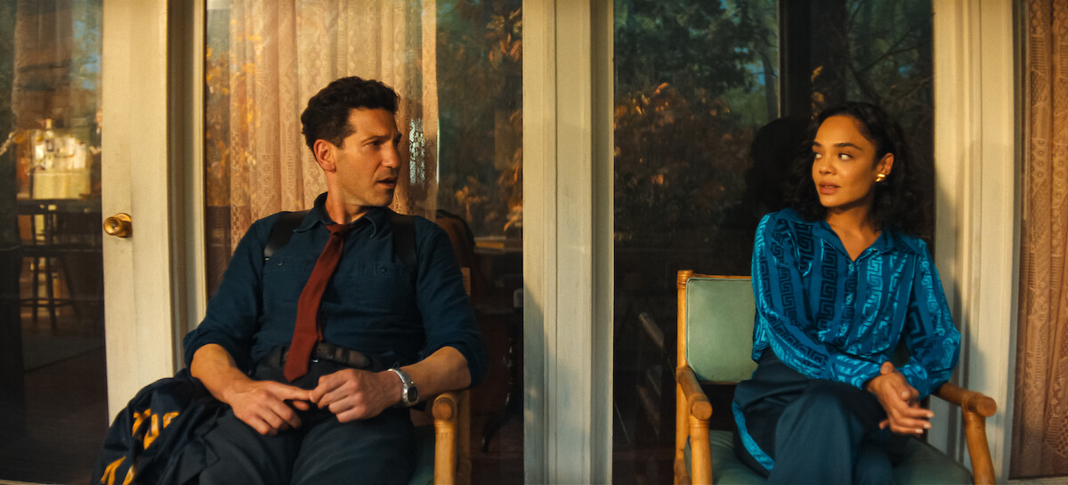 A man and woman sit on chairs outside near glass doors, engaged in a serious conversation.