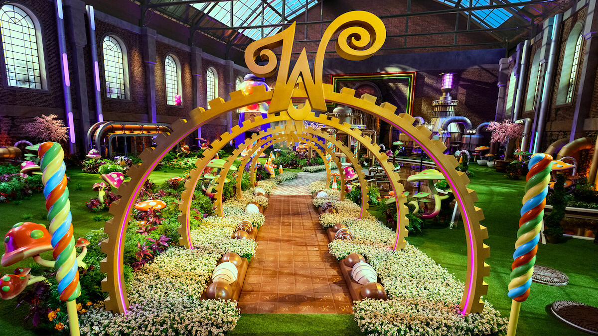 A whimsical candy-themed garden inside a large factory with colorful sweets, oversized mushrooms, lollipops, golden arches, flowers, and a large "W" centerpiece under a glass roof, creating a magical, playful atmosphere.
