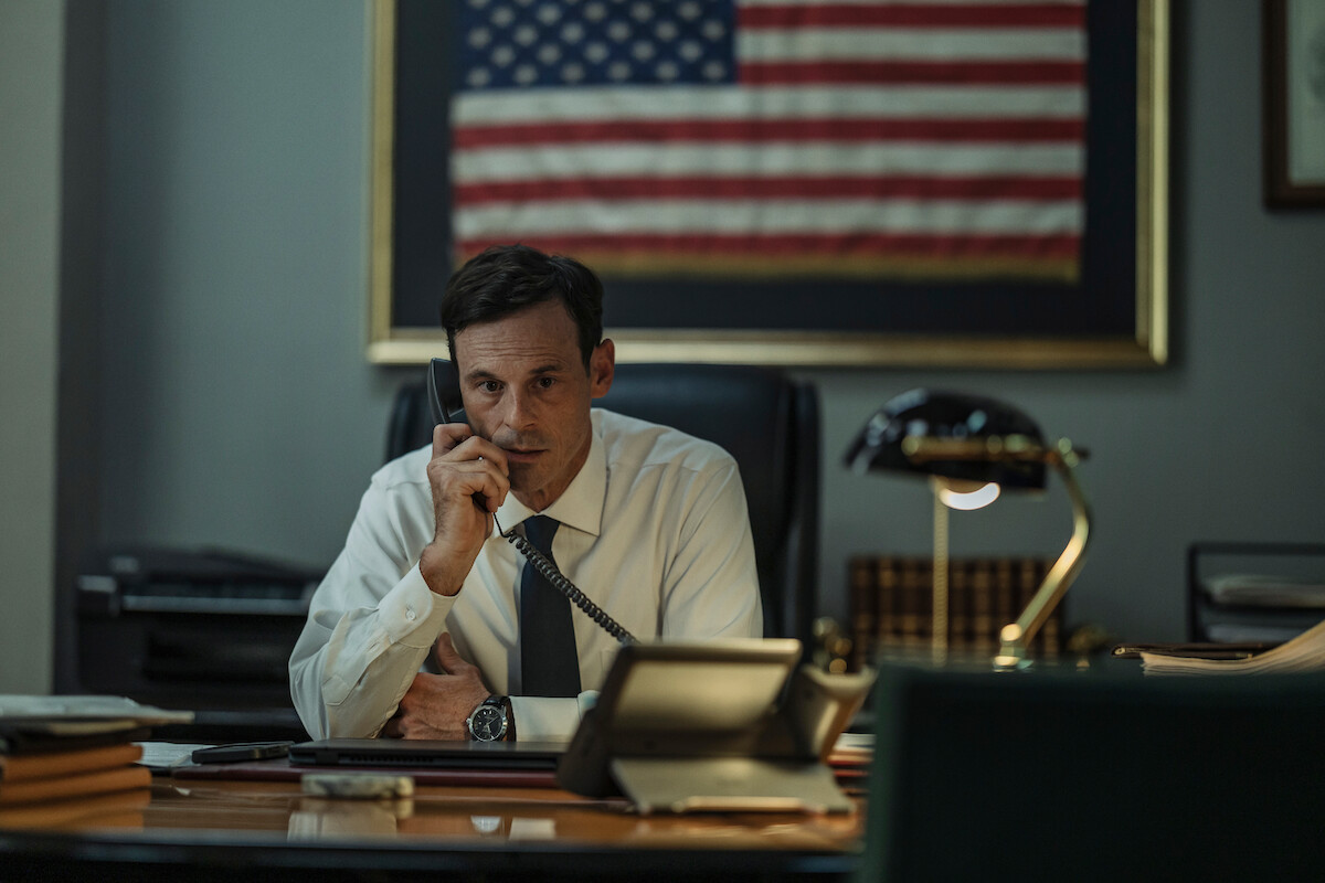 Man in formal attire sitting at a desk, talking on a corded phone in an office. Behind him is a large framed American flag on the wall, and there are office supplies and a lamp on the desk.