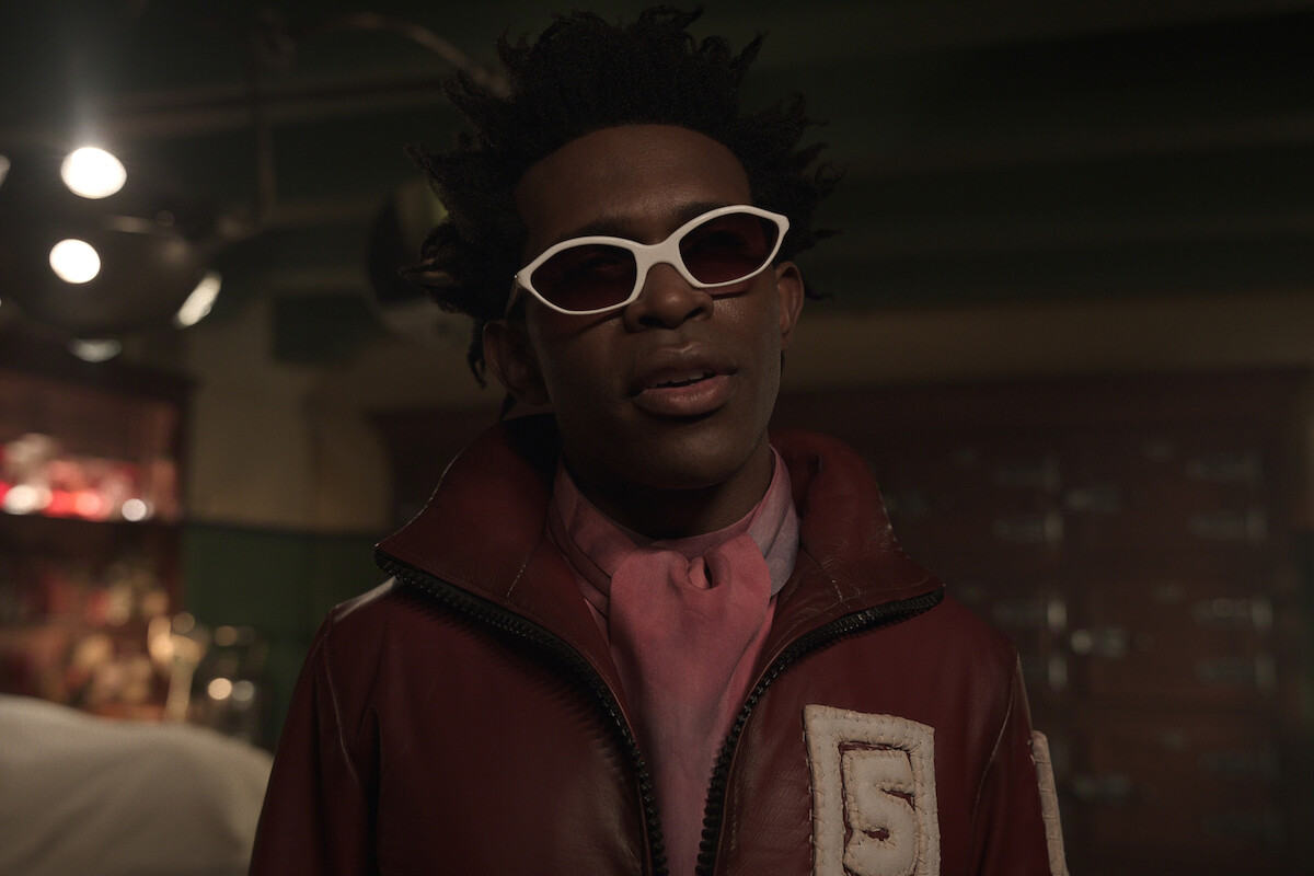 Person wearing white sunglasses, red jacket with number 5 patch, and pink scarf stands indoors in a dimly lit room with green walls and vintage decor.