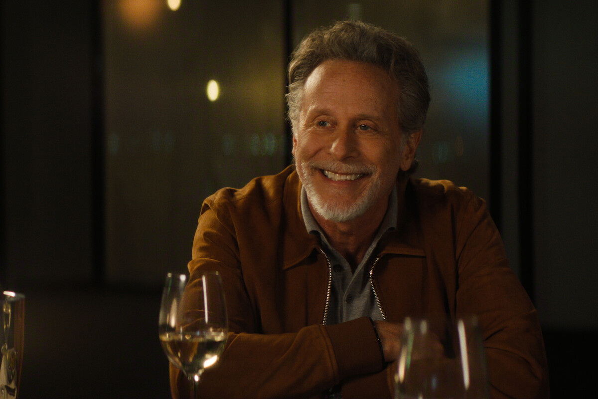 Smiling older man with gray hair and beard in a brown jacket sits at a dimly lit restaurant table with wine glasses, blurred city lights in background, creating a warm, relaxed atmosphere.