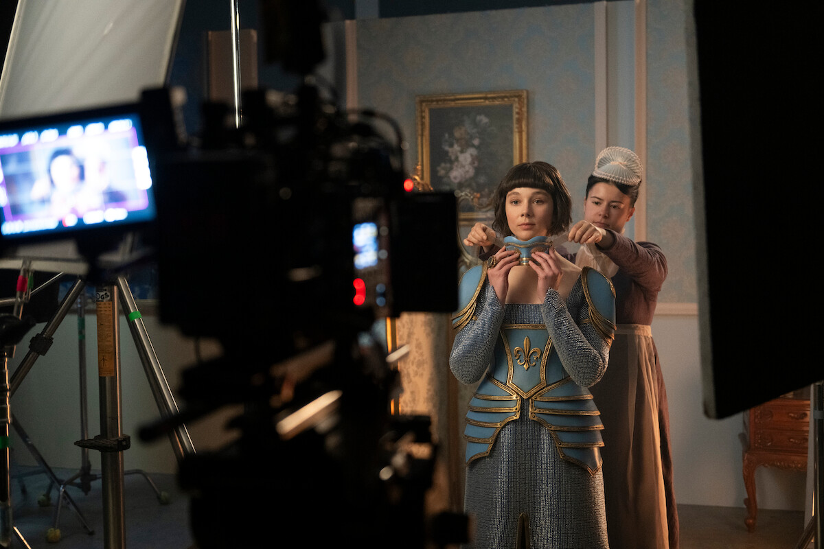 Em um set de filmagem, uma mulher recebe ajuda de outra para ajustar o figurino, cercada por refletores e equipamentos de câmera em uma sala antiga e ornamentada.
