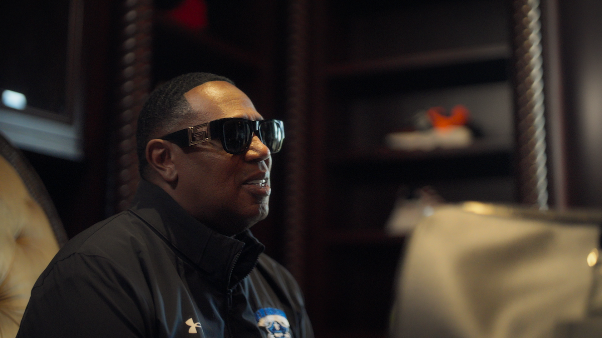 Man wearing sunglasses and a dark jacket sits in a dimly lit room with wooden shelves, a chair, and some shoes in the background. The mood is relaxed and the setting appears to be an office or study.