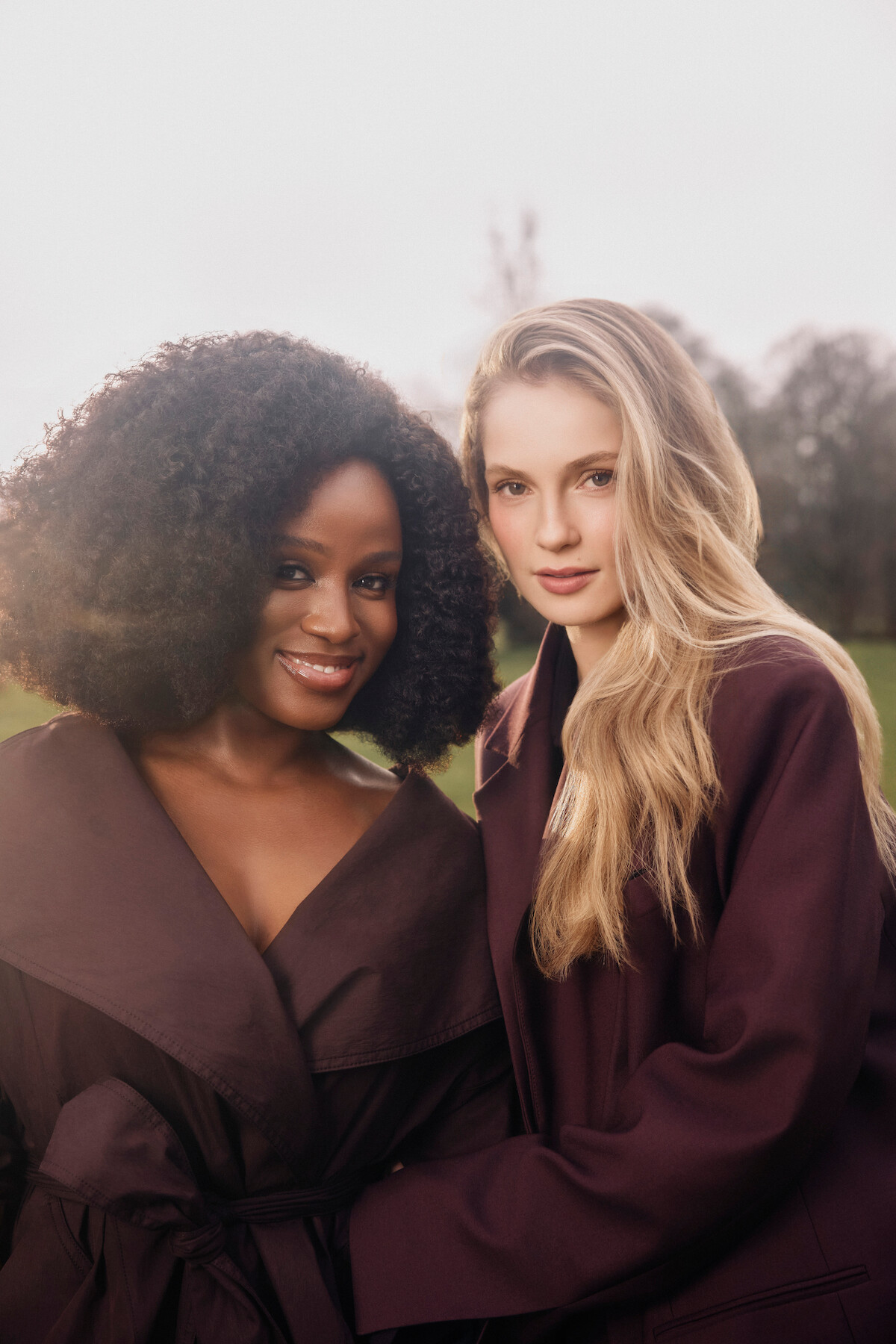 Two women in dark coats standing close together outdoors, smiling softly, with trees and a cloudy sky in the background, natural light creating a gentle and warm atmosphere.