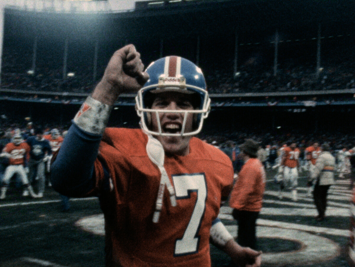 John Elway celebrates passionately on field, surrounded by teammates and fans in a stadium, capturing the excitement and energy of a game day victory.