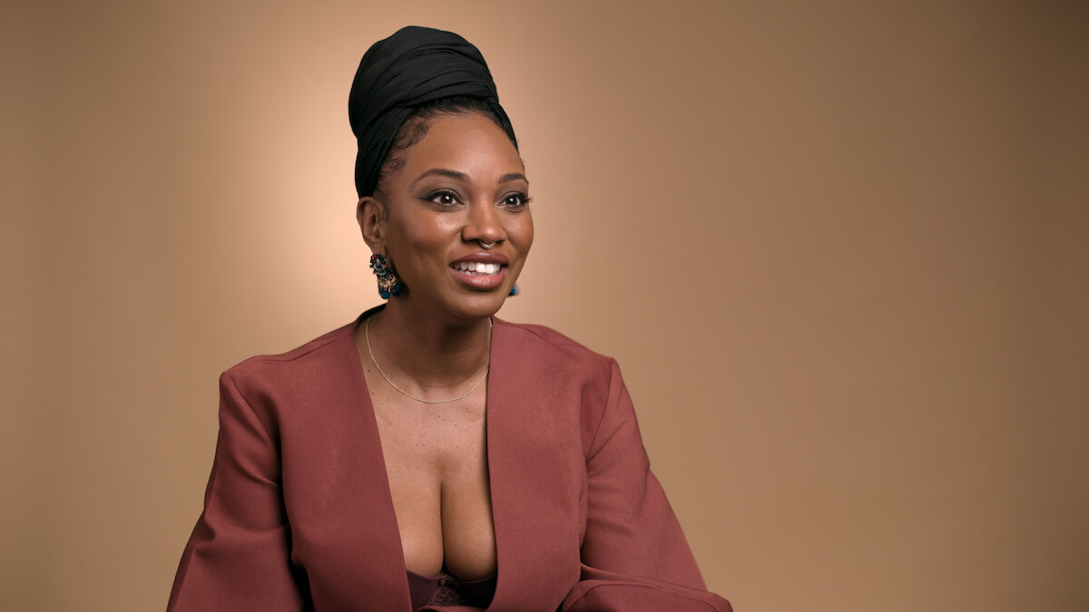 Smiling woman with a headwrap and deep V-neck mauve blazer, wearing earrings, sitting in front of a plain beige background, studio lighting, posed for a portrait.