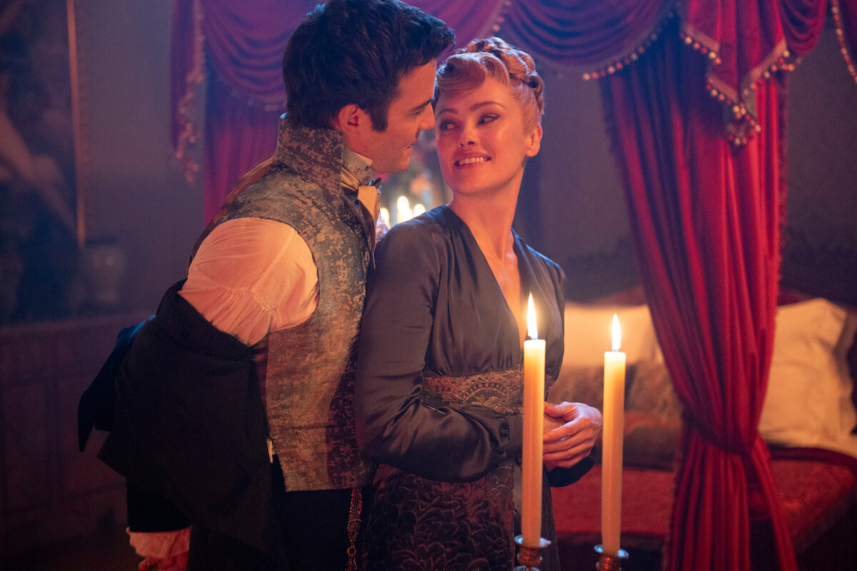 A couple dressed in historical costumes stands close together in a lavish, dimly lit room with red curtains and candles, creating a romantic and intimate atmosphere.