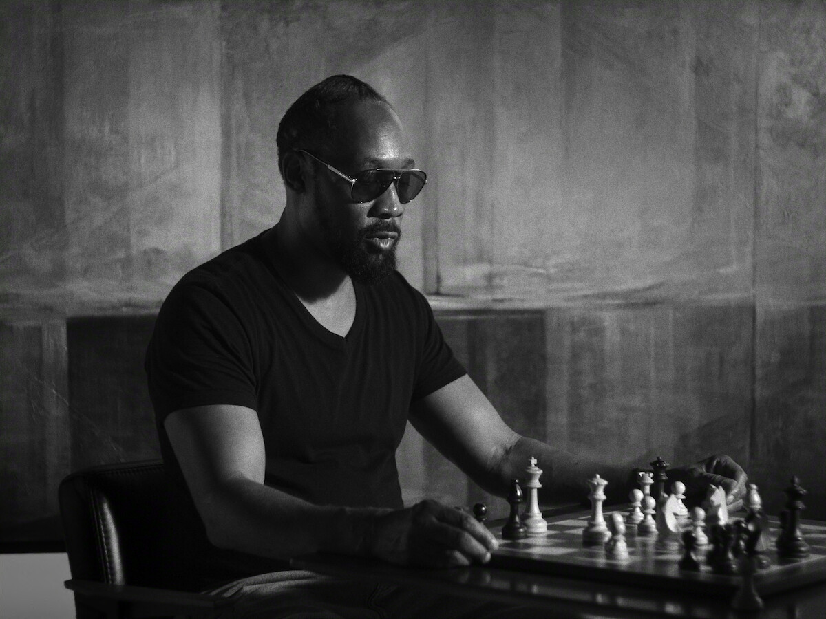 Man wearing sunglasses and a dark t-shirt sits at a table concentrating on a chess game in a dimly lit, modern room with abstract patterned walls. The scene is in black and white, evoking a thoughtful, serious mood.