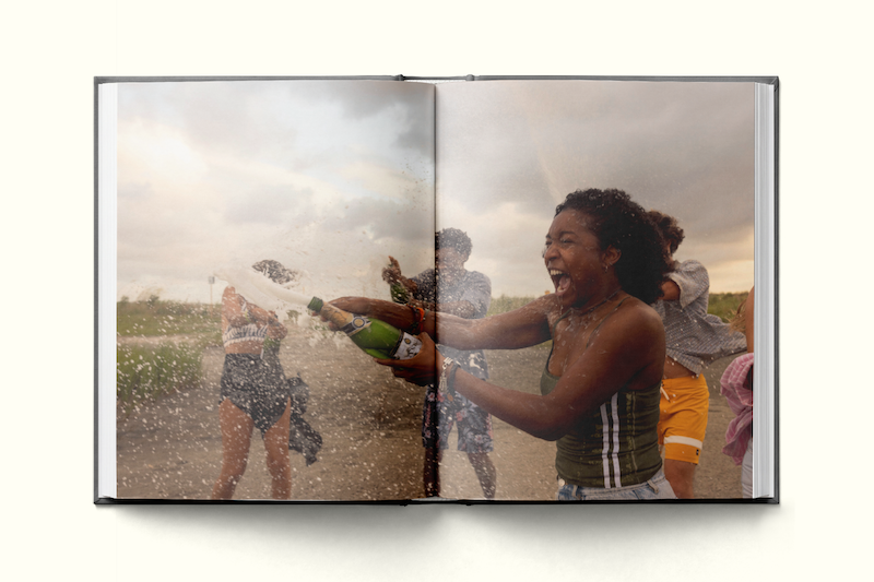 A joyful group of young people outdoors, one woman in front sprays champagne, laughing, with friends in the background, grassy field, cloudy sky, captured in an open photo book.