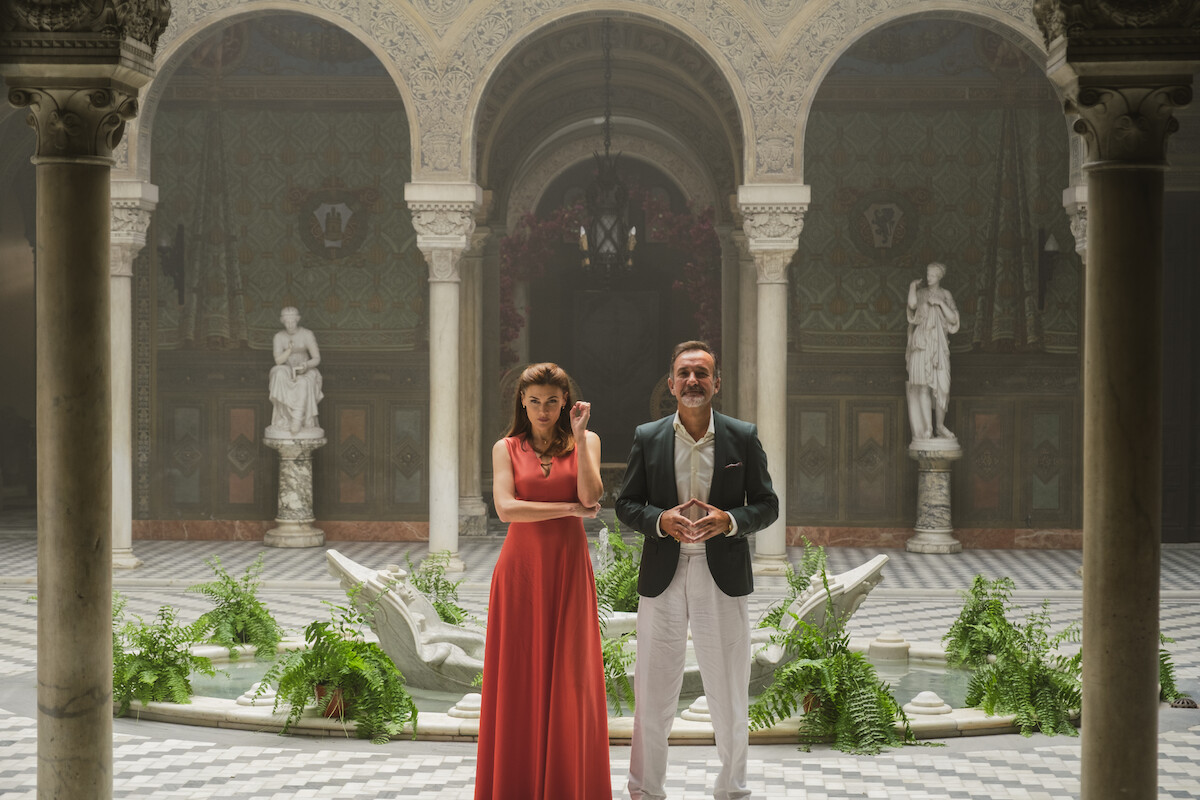 A woman in a red dress and a man in a suit stand in an ornate courtyard with marble columns, statues, a patterned floor, plants, and arched ceilings, creating a formal and elegant setting.