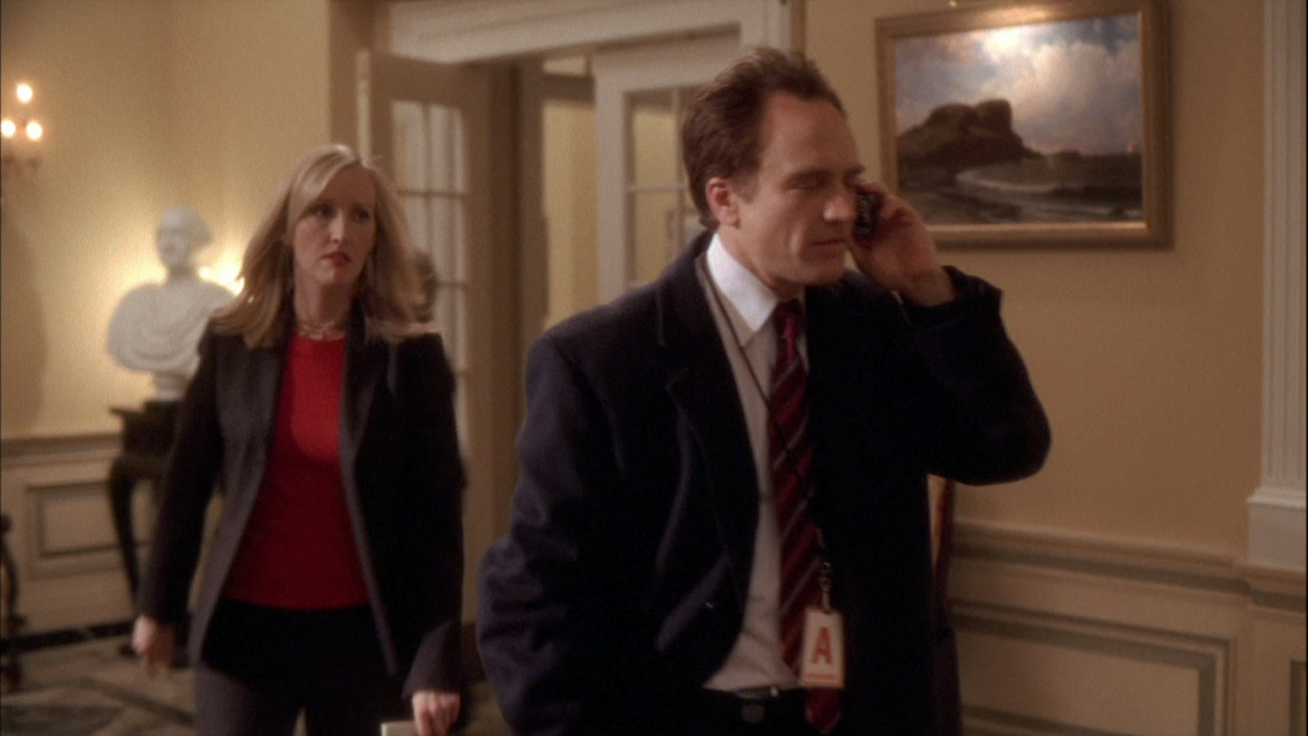 Man in suit talking on phone walks through elegant office hallway with a woman following; art, bust, and classic decor visible in background.