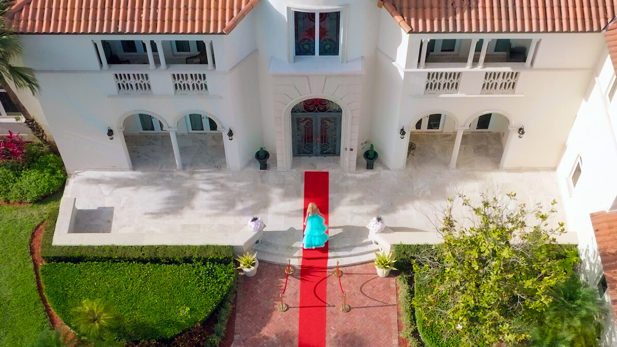 A person in a blue dress walks up a red carpet towards the entrance of a large, elegant white mansion surrounded by manicured gardens, creating a luxurious and celebratory atmosphere.