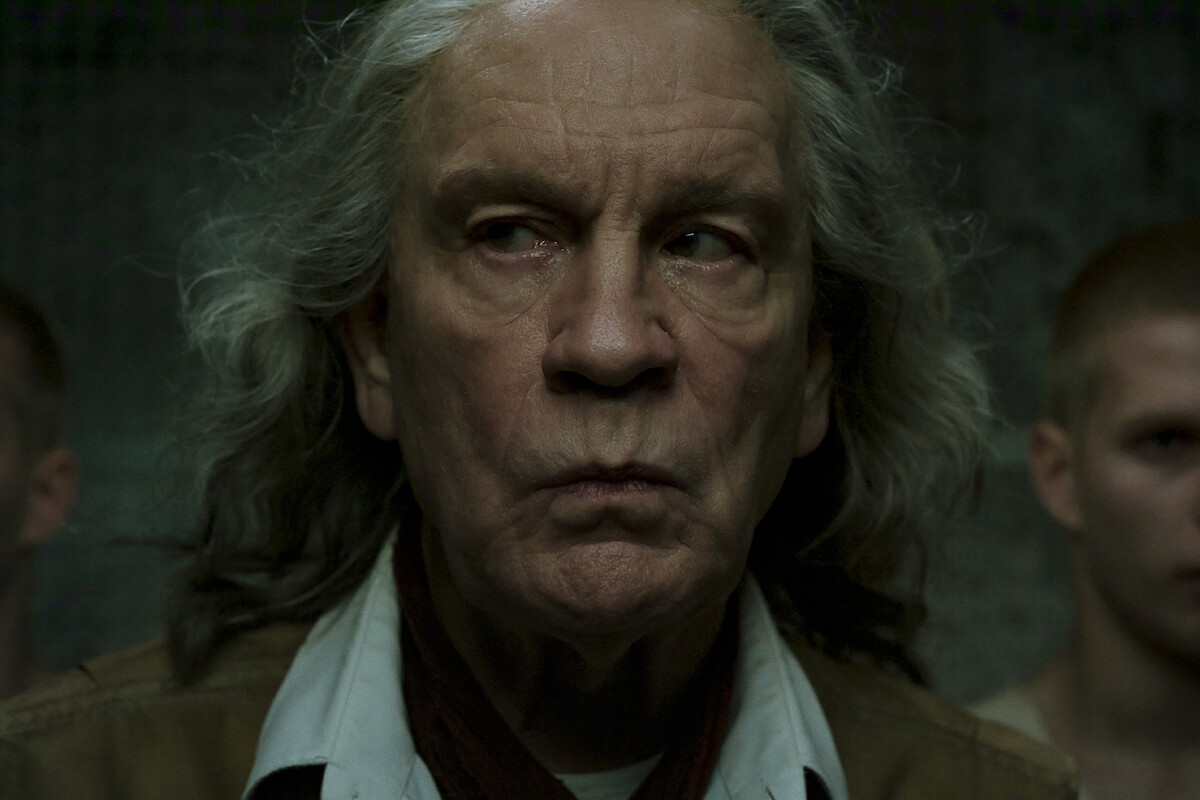 Older man with long gray hair and serious expression stands indoors in dim lighting, with two younger people with short hair blurred in the background.